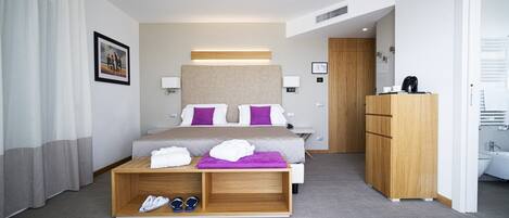 A modern hotel room with a large bed, a wooden desk, a wardrobe, and a bathroom visible through an open door.