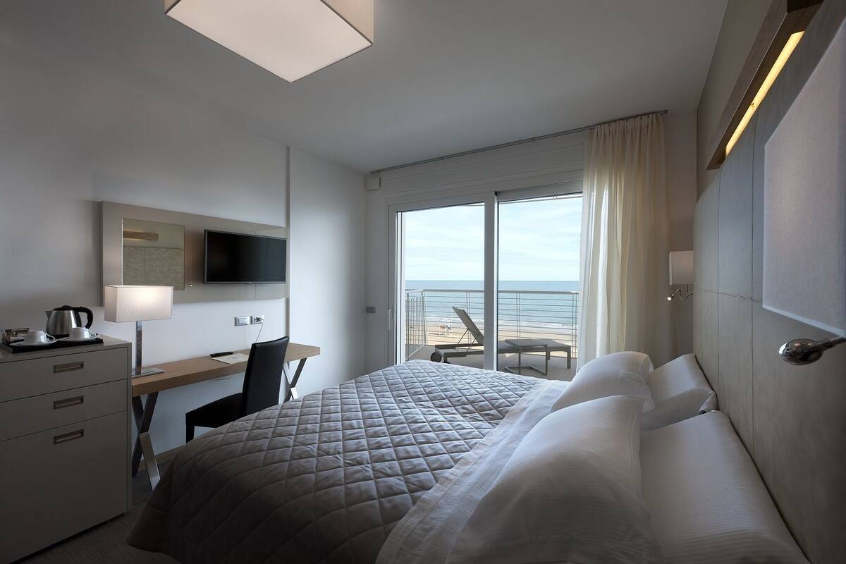 Ett hotellrum med en säng, ett skrivbord med en stol, en tv och utsikt över stranden.