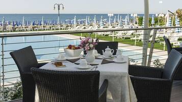 A dining area with a view of the sea, featuring wicker chairs, a table set with white tableware, and a flower vase.