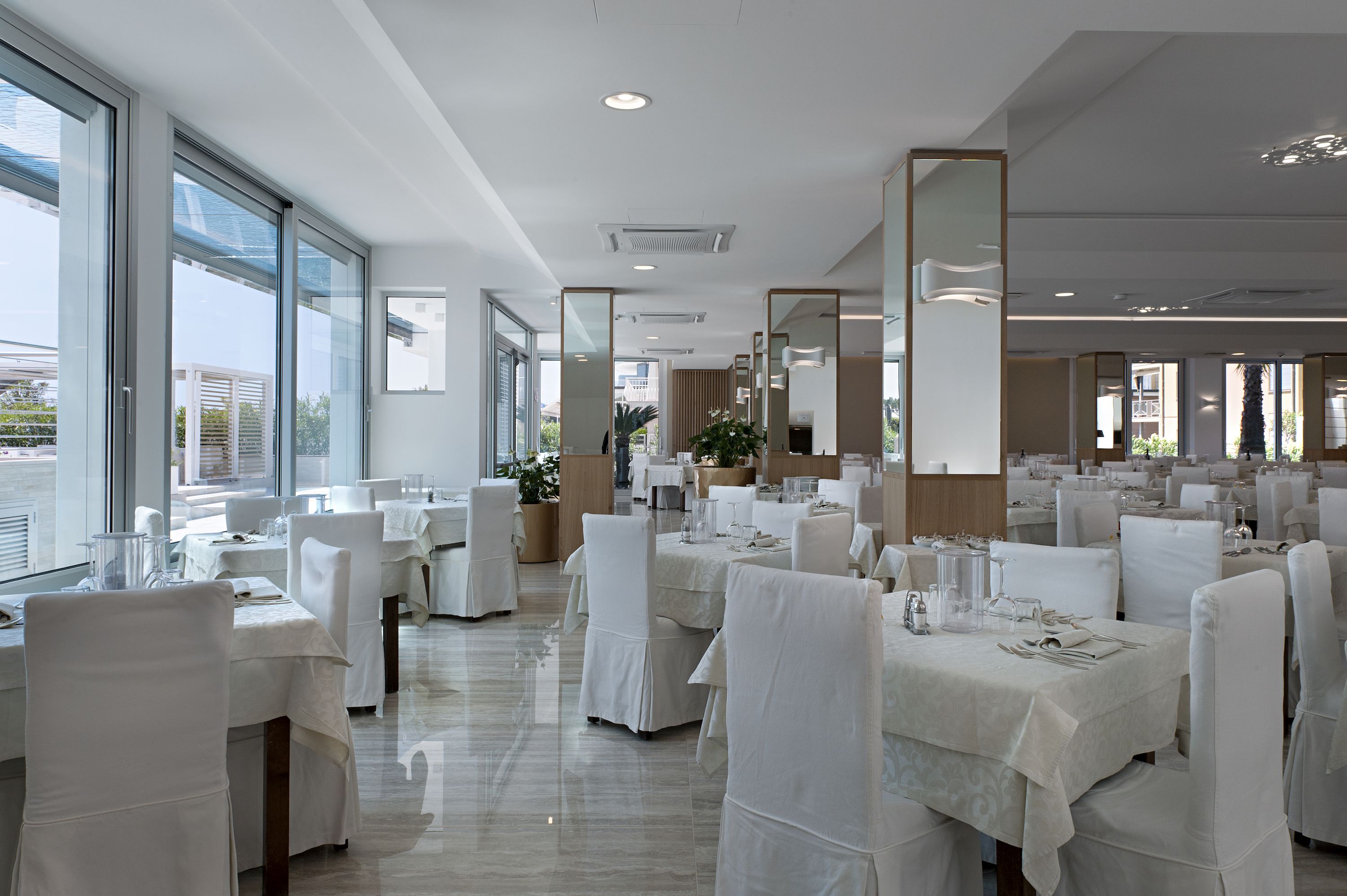 Ein geräumiges Restaurant mit hohen Decken, großen Fenstern und moderner Einrichtung. Die Tische sind mit weißen Tischdecken ordentlich gedeckt und mit Stühlen bestückt.