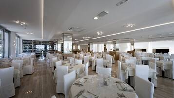 A spacious banquet hall with round tables set for dining, white chairs, and a wine rack in the background.