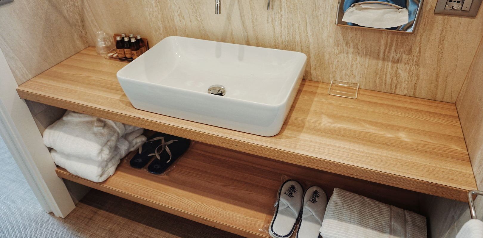 A bathroom with a white rectangular sink on a wooden vanity, a towel rack above the sink, and a shelf below holding folded towels and a pair of slippers.