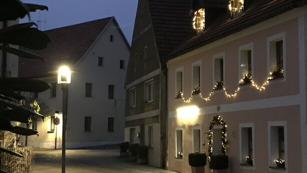 A street scene at dusk with buildings featuring traditional architecture, including a pink building with string lights and a white building with a street lamp. - Haus am Markt (Königstein)