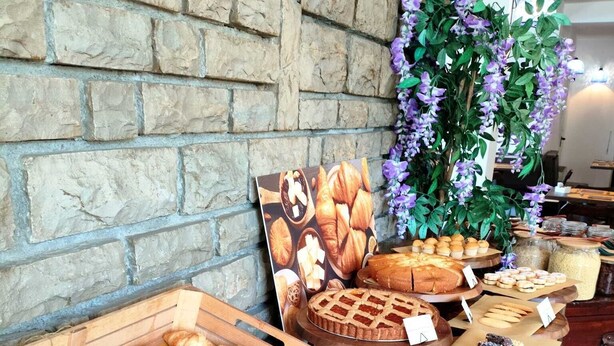 A bakery display with various baked goods in wooden crates, a wall with a brick pattern, and a decorative plant with purple flowers.