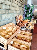 Una vitrina de panadería con diversas opciones de panadería en cajas de madera, una pared con diseño de ladrillo y una planta decorativa con flores púrpuras.