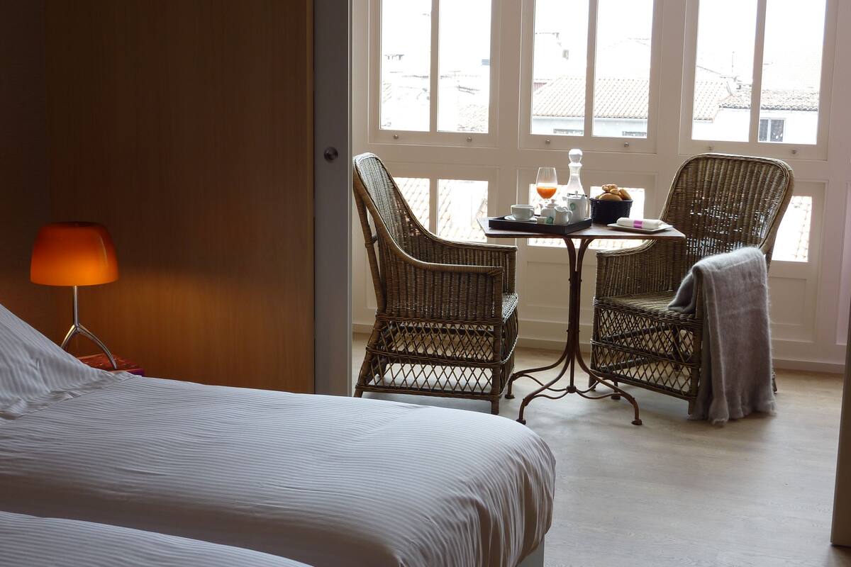 A hotel room with a bed, a table with chairs, a lamp, and a window with a view of buildings.