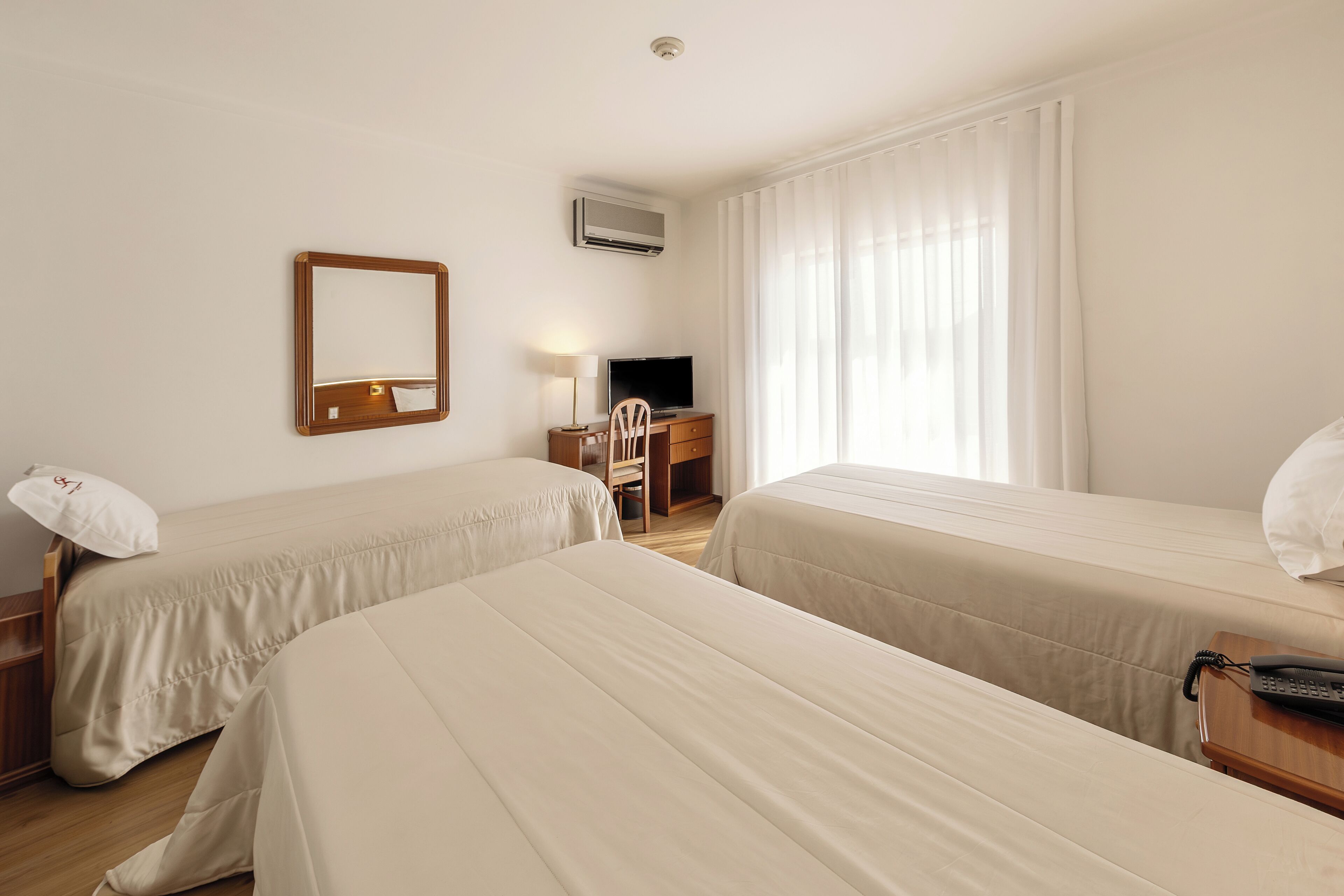 A hotel room with two beds, a desk with a chair, a television, an air conditioning unit, and a telephone.