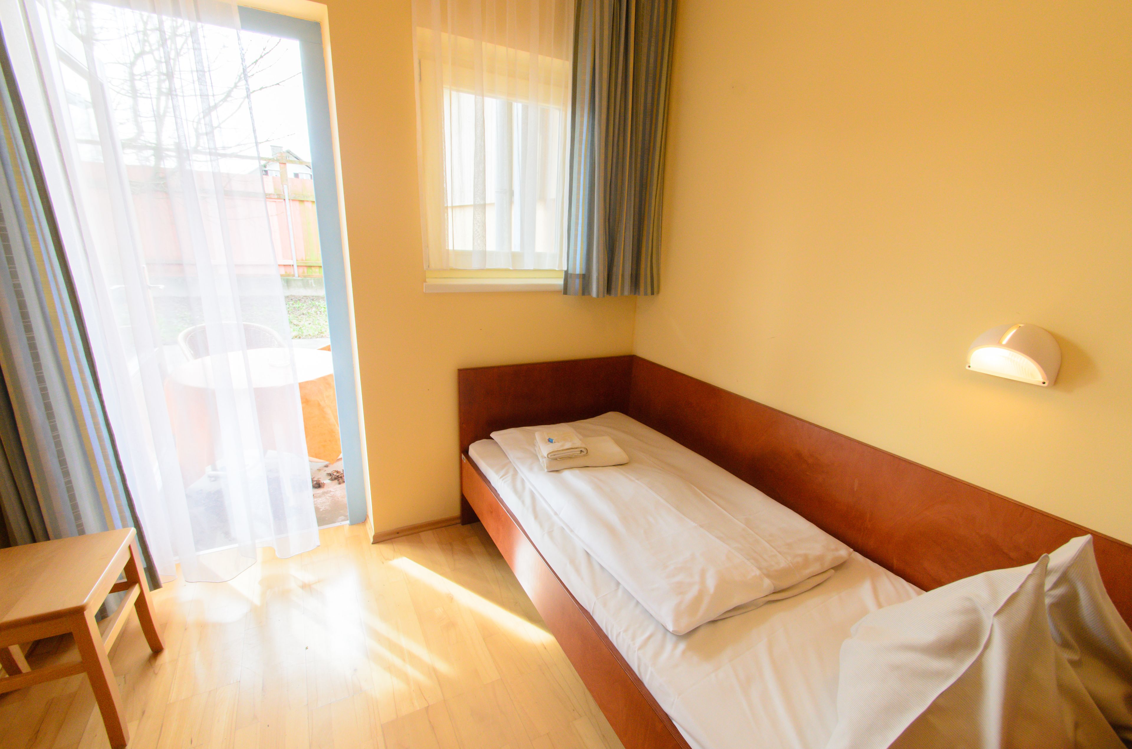 Een enkel bed met een houten hoofdeinde, een nachtkastje en een raam met transparante gordijnen.