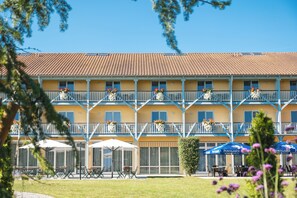 A hotel with a yellow facade, multiple balconies with flower planters, outdoor seating with umbrellas, and a clear blue sky. - JUFA Hotel Vulkanland Sport- Resort (Gnas)