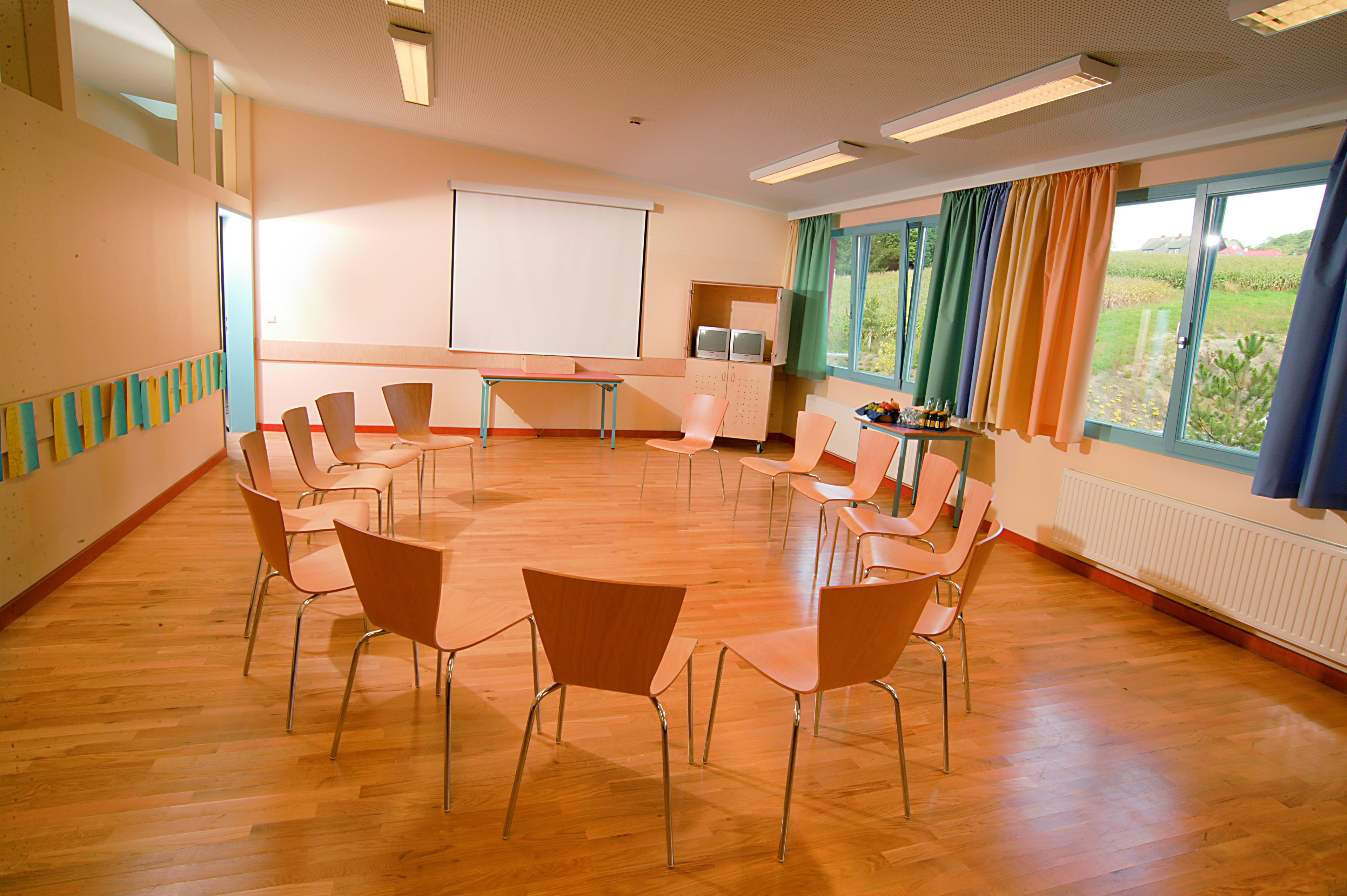 Una sala de conferencias con una mesa redonda, sillas, una pantalla de proyector y ventanas con cortinas.