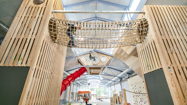 Espaço de escalada indoor com estruturas de madeira, um escorrega vermelho e paredes de escalada.