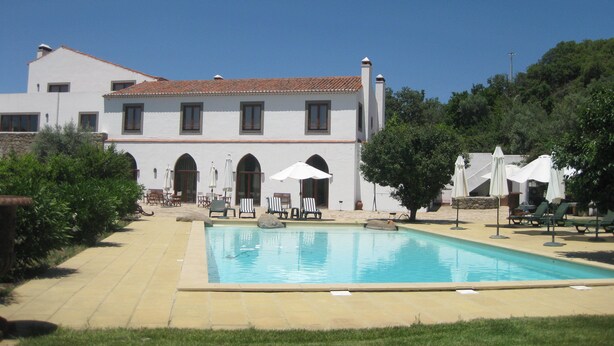 Un espace piscine avec des chaises longues et des parasols, un bâtiment blanc doté d’un toit en tuiles et un ciel d’un bleu limpide.