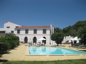 A pool area with lounge chairs and umbrellas, a white building with a tiled roof, and a clear blue sky.