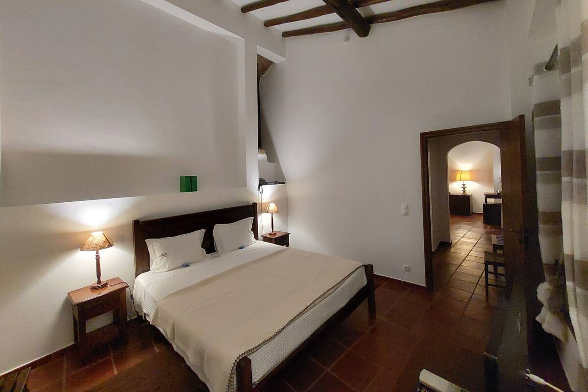 A bedroom with a large bed, wooden beams on the ceiling, and a small table with a lamp.