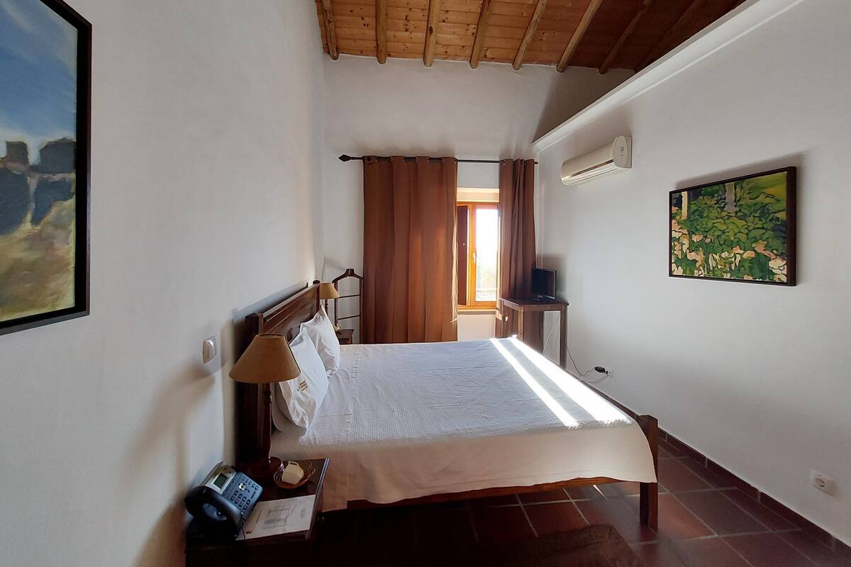 A bedroom with a wooden ceiling, a bed with white linens, a bedside lamp, a telephone, and a painting on the wall.