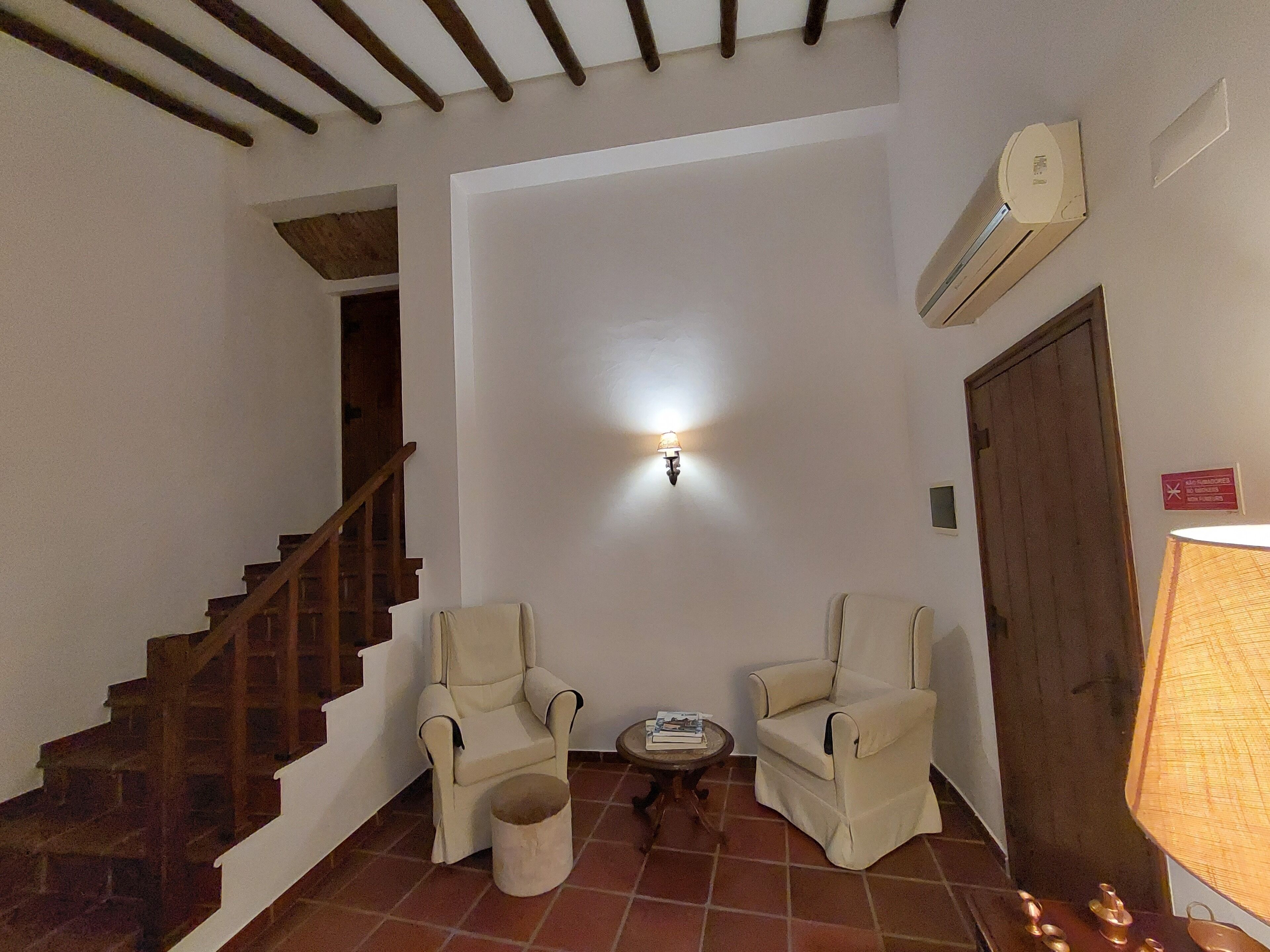 Una habitación con una escalera, dos sillones, una mesita con libros, una lámpara y un equipo de aire acondicionado.