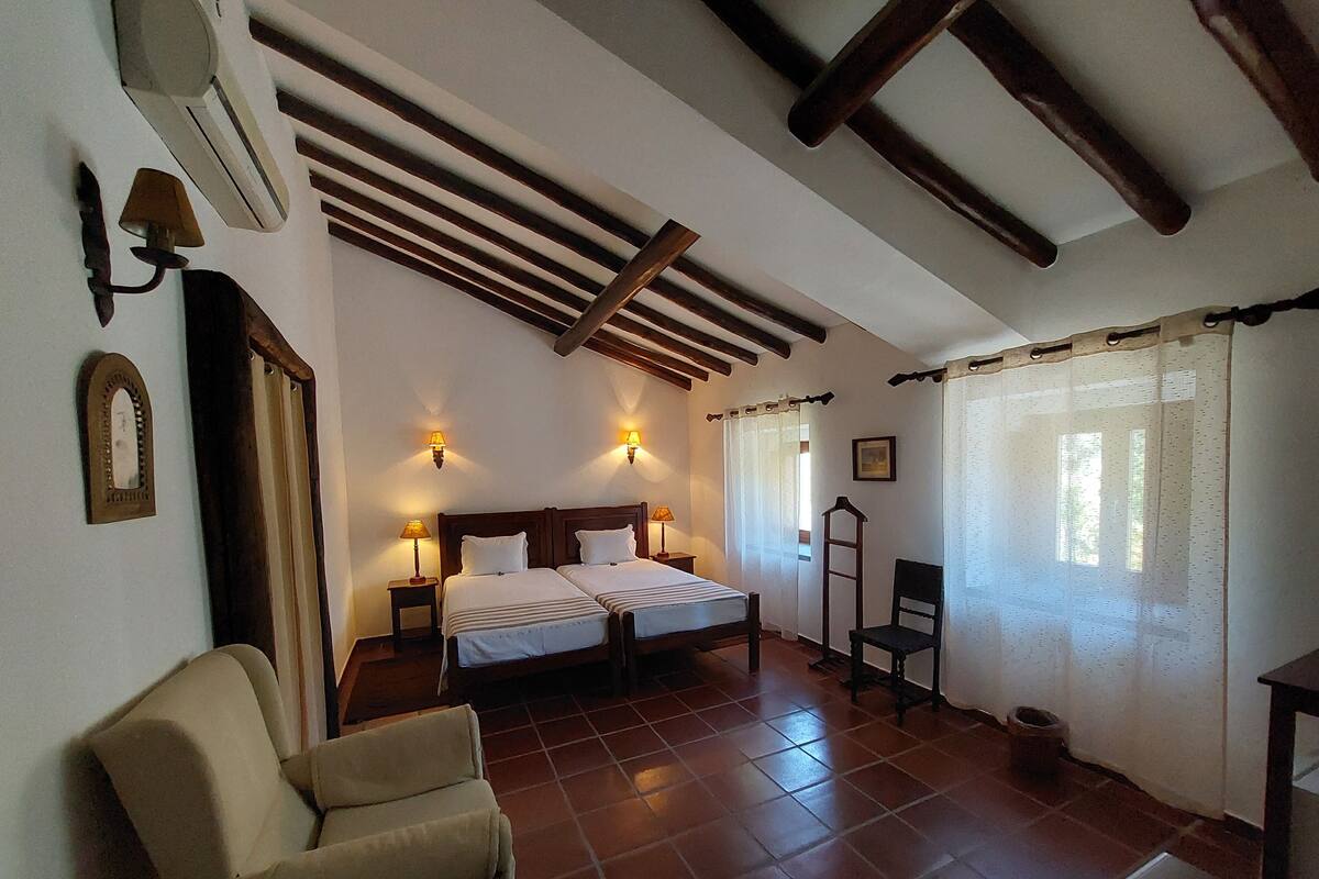 A bedroom with two beds, a chair, a wooden ceiling, and a tiled floor.