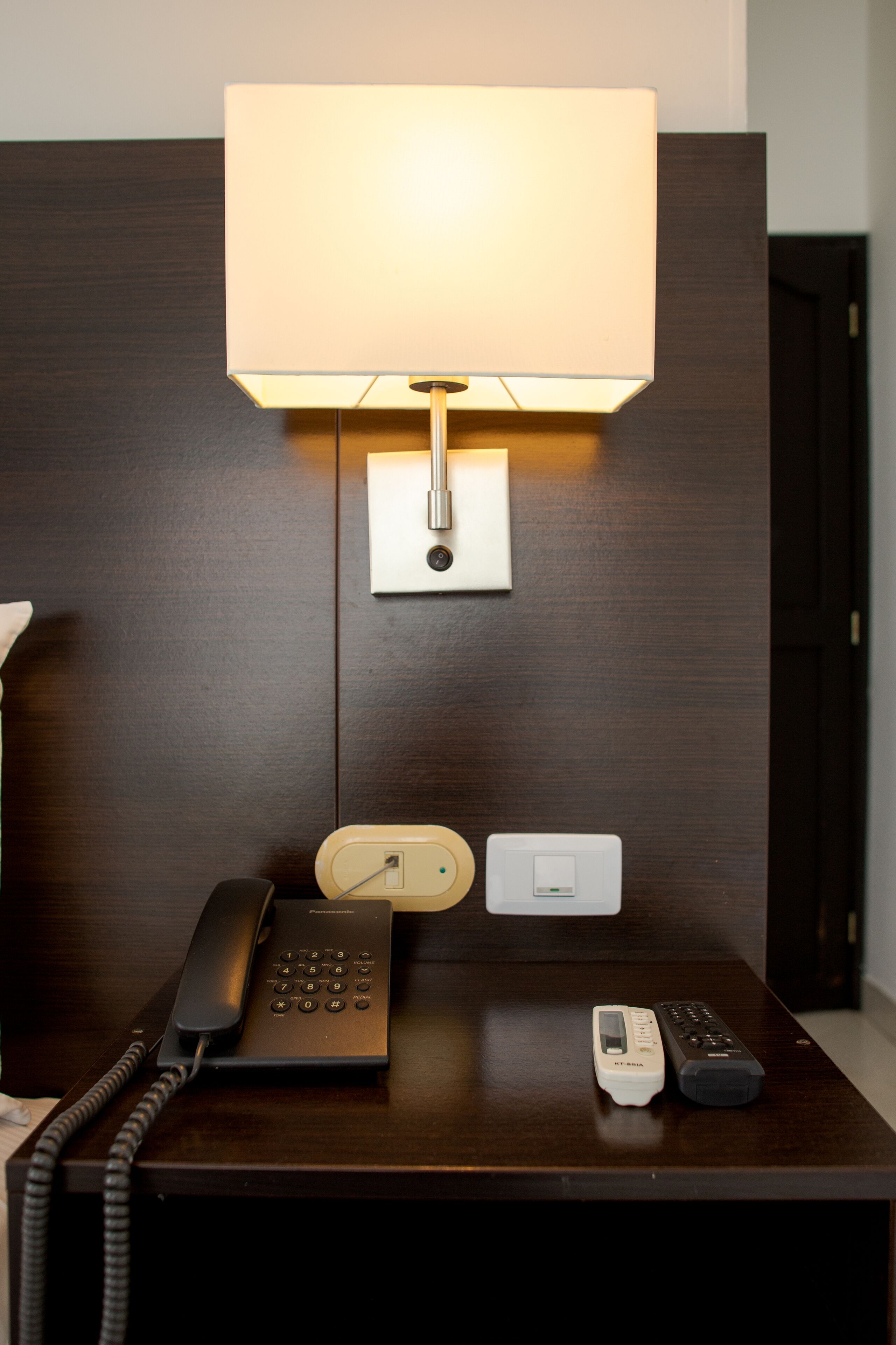 Una habitación de hotel con escritorio, lámpara, teléfono y control remoto.