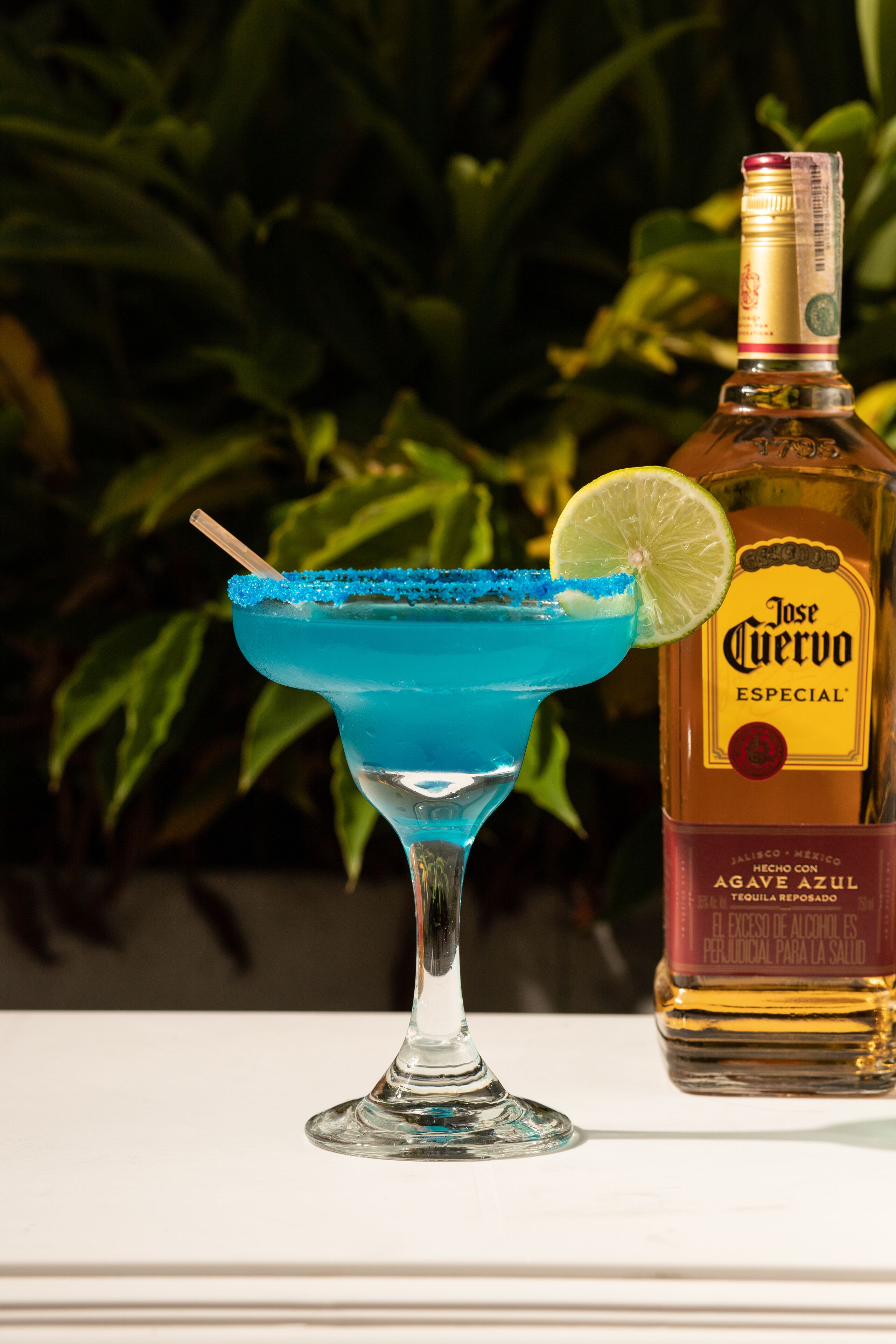 A blue cocktail with a lime garnish and a straw, next to a bottle of Jose Cuervo Especial tequila.
