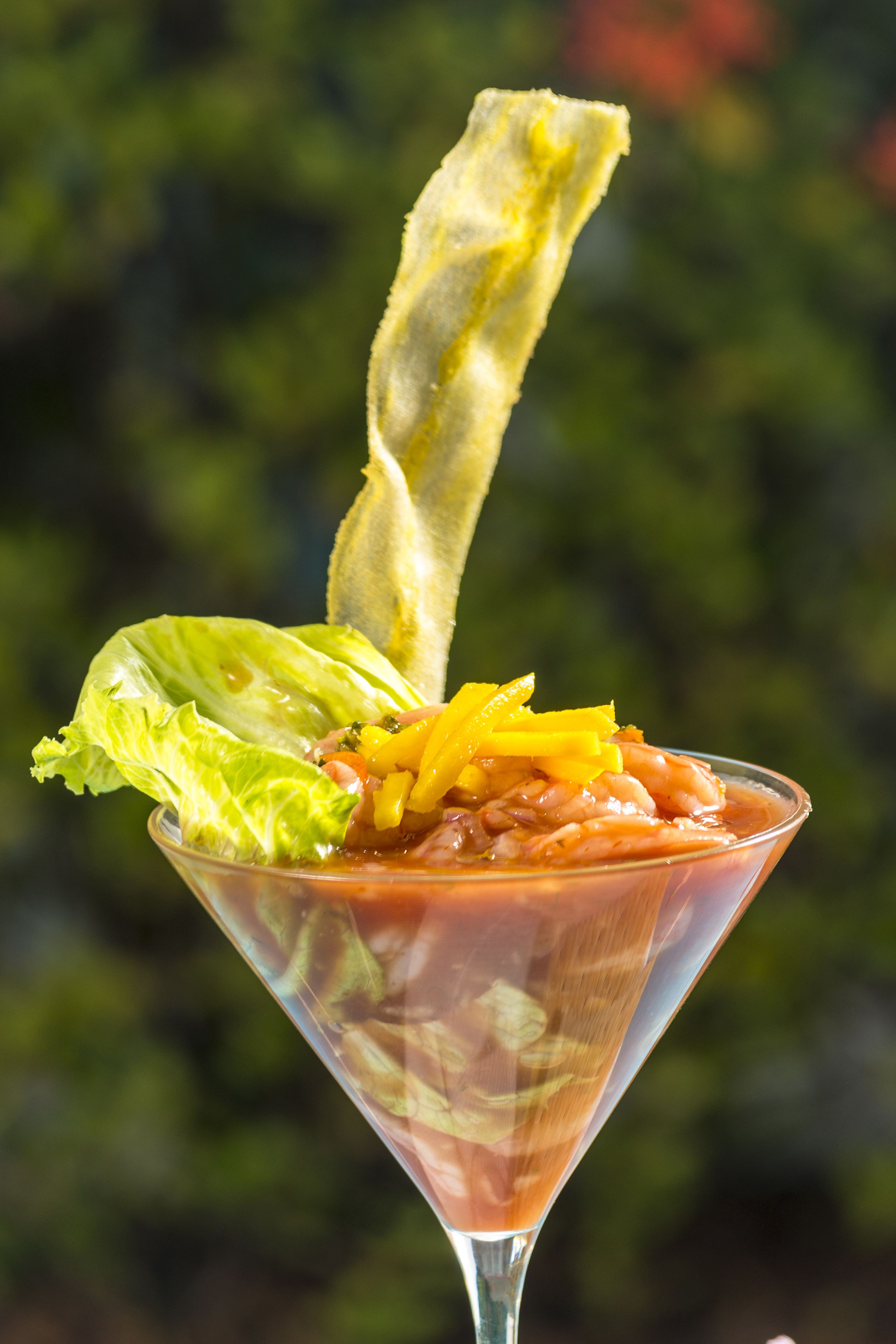 Un verre à martini, garni de feuilles de laitue et de tranches d’orange, contenant un liquide transparent, peut-être un cocktail, le tout sur un fond vert flou.