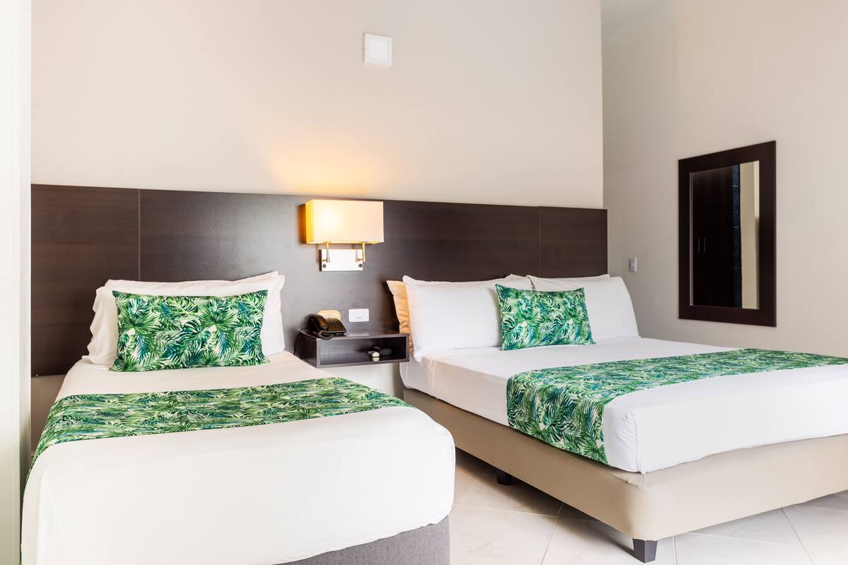 A hotel room with two beds, each with a green leaf-patterned bedspread and matching pillows. There is a nightstand with a lamp between the beds.