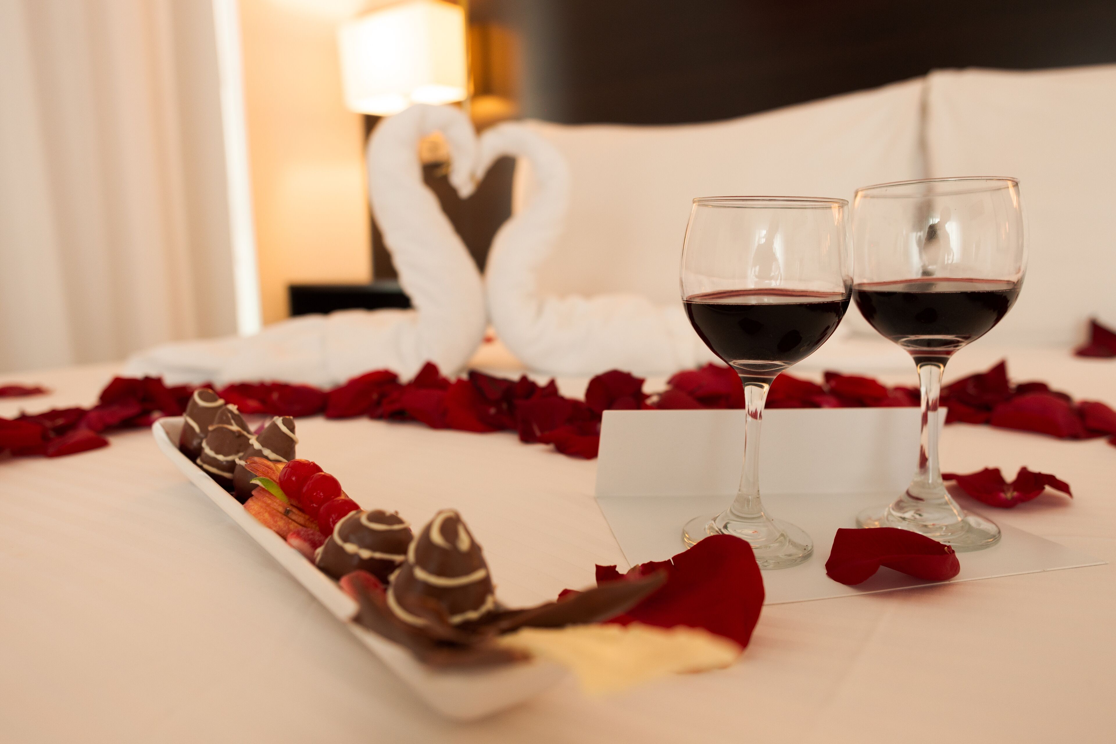 Una cama con pétalos de rosa, dos copas de vino y una bandeja con postres de chocolate.