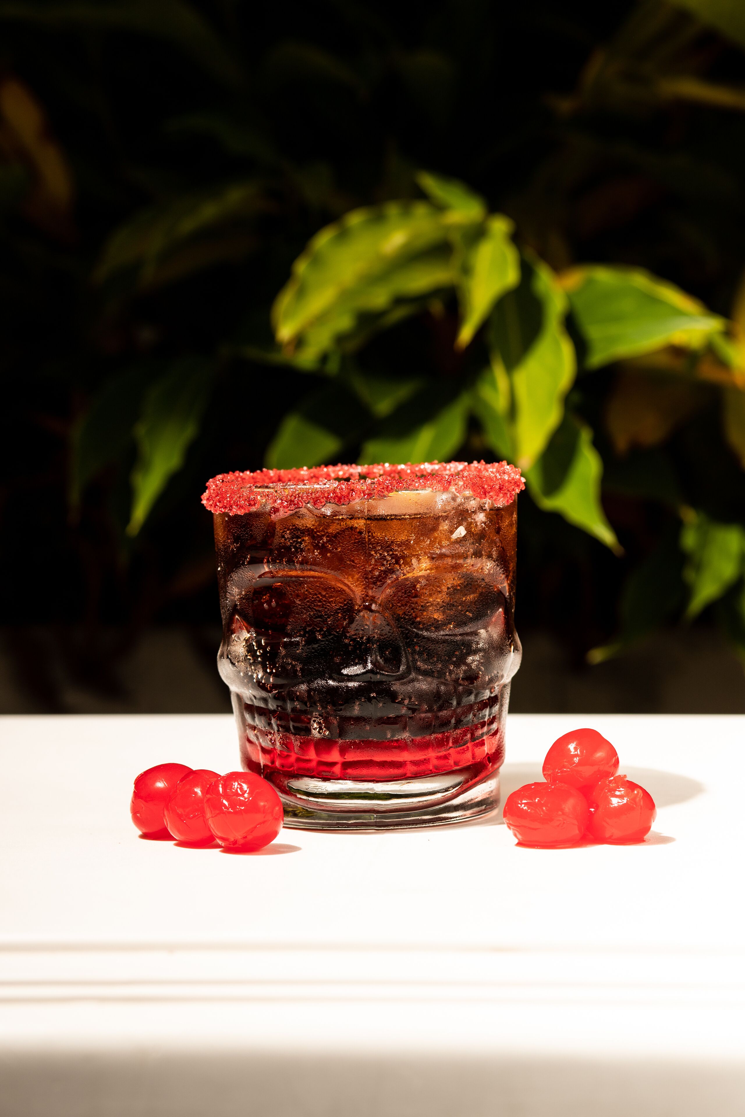 Ein Glas mit salzigem Rand, Eiswürfeln und roten Fruchtstücken auf einer weißen Oberfläche, im Hintergrund grüne Blätter.