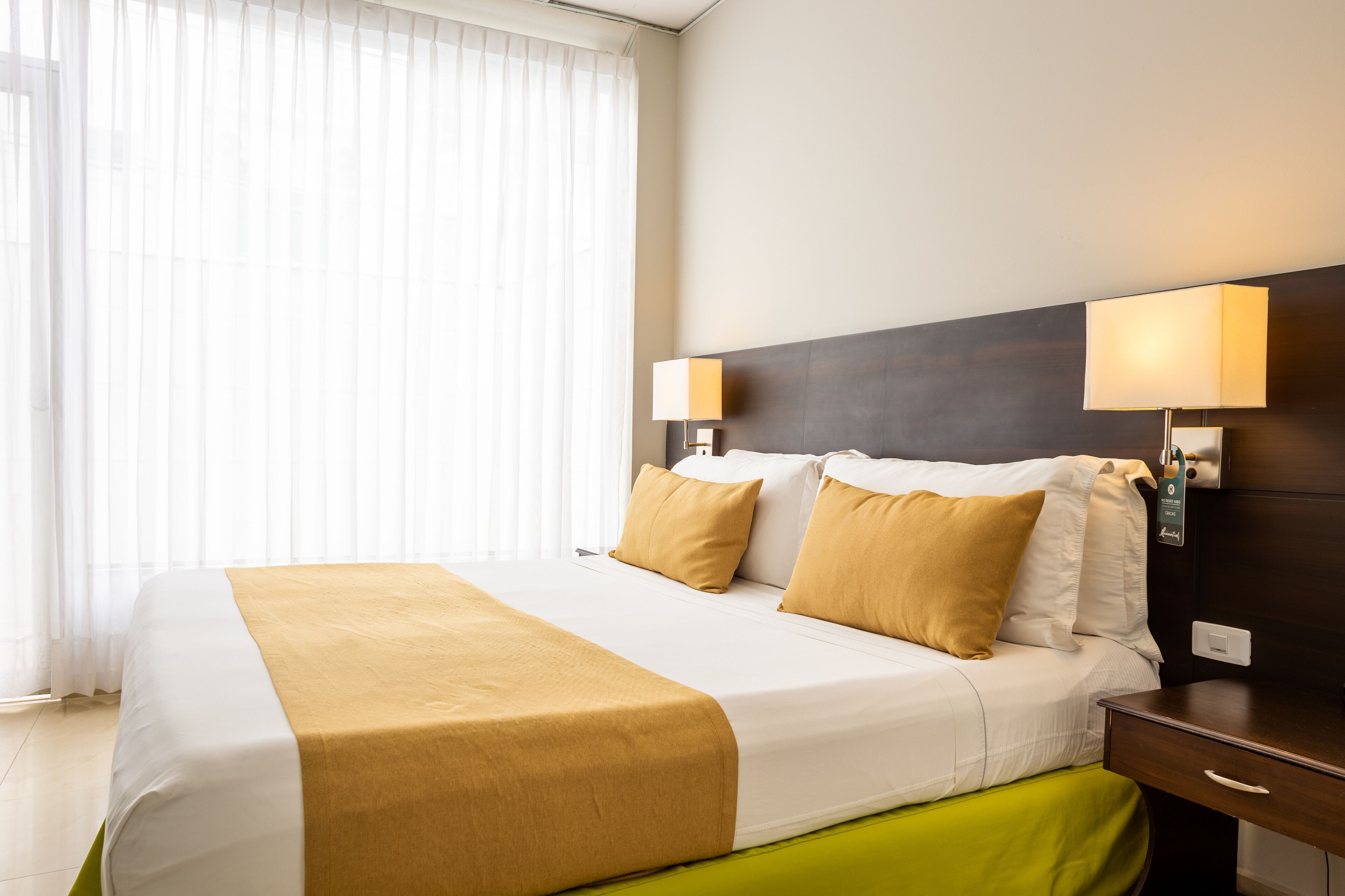Habitación de hotel con una cama grande, dos lámparas de noche, una mesita de noche y un ventanal con cortinas translúcidas.