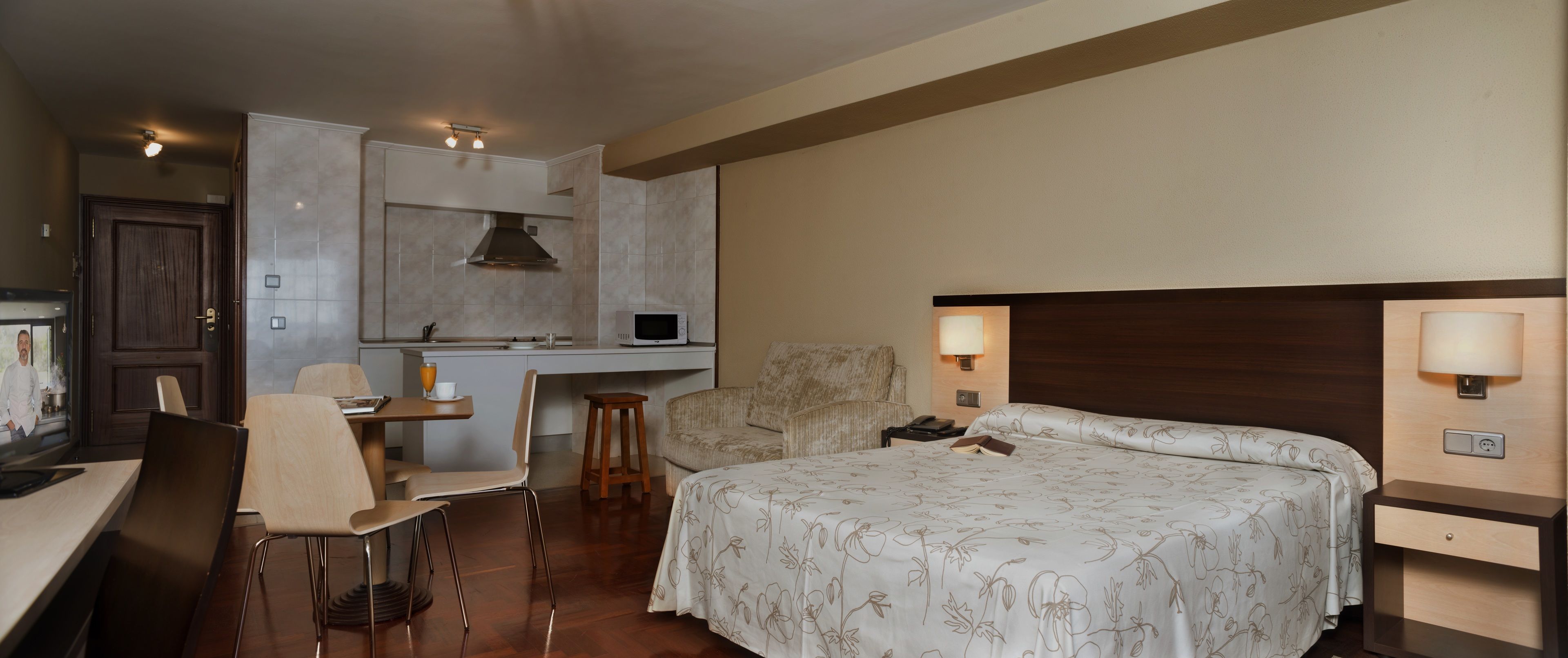 A hotel room with a bed, a desk, a chair, a small stool, a TV, and a microwave.