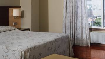 Una habitación de hotel con cama, mesita de noche con lámpara, ventana con cortinas y vista a edificios.