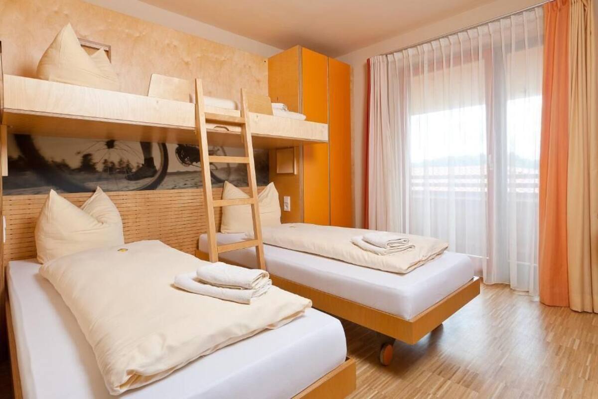 Una habitación con dos camas, una litera, suelo de madera y un ventanal con cortinas.