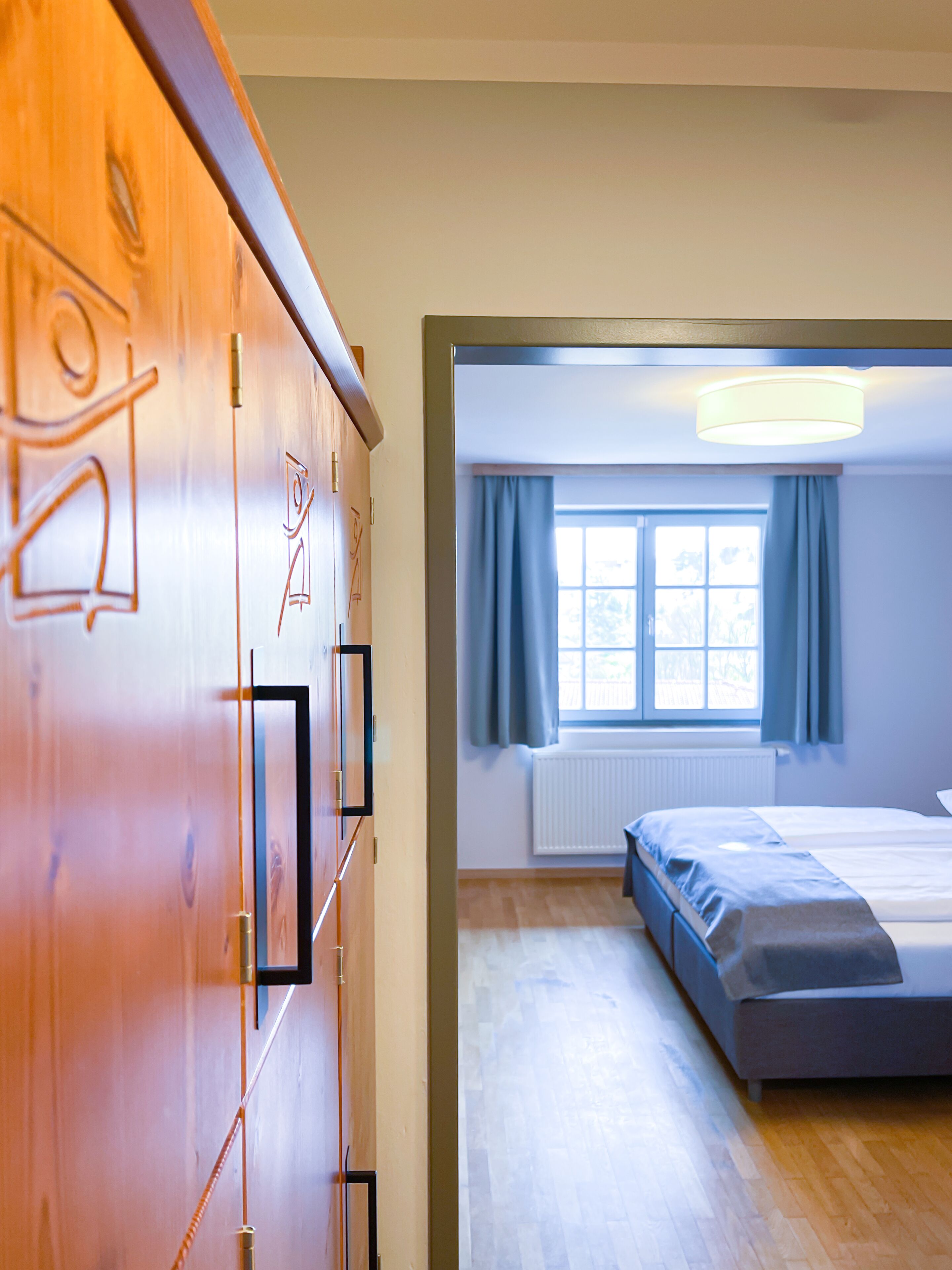 A bedroom with a bed, a window with curtains, a radiator, and a wooden wardrobe with a mirror.