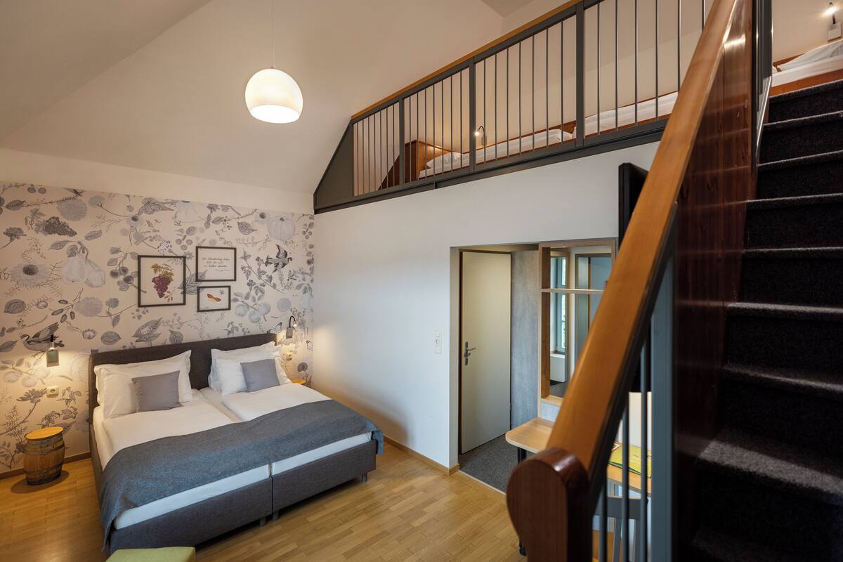 Un dormitorio con cama, piso de madera, una escalera y una pared con papel tapiz floral.