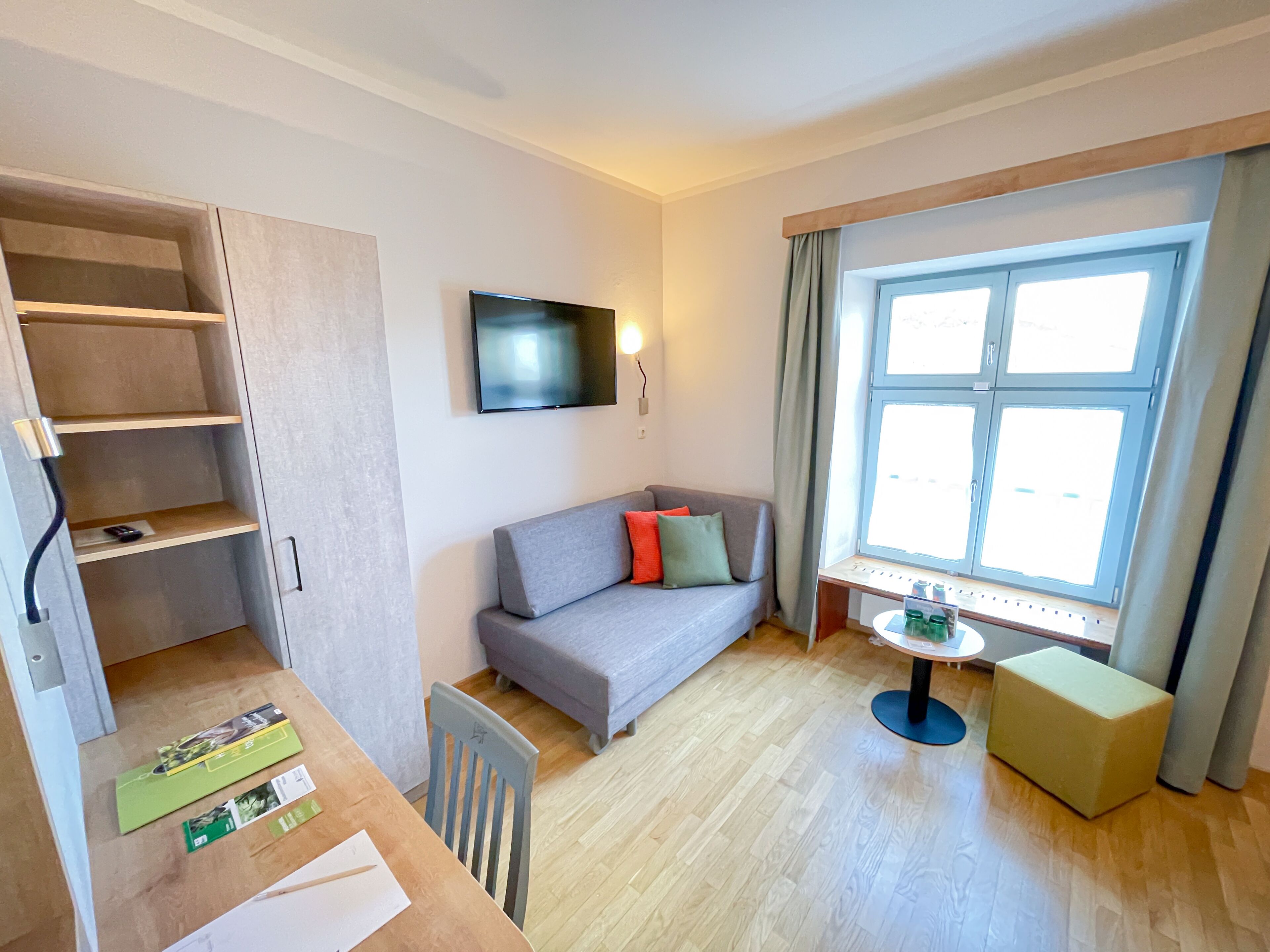 Un espacio habitable pequeño con un sofá gris, una mesita, un televisor montado en la pared y una ventana con cortinas.