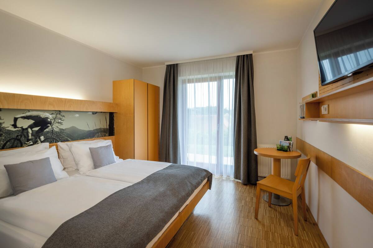 Habitación de hotel con cama, una mesita, una silla, televisor y ventana con cortinas.