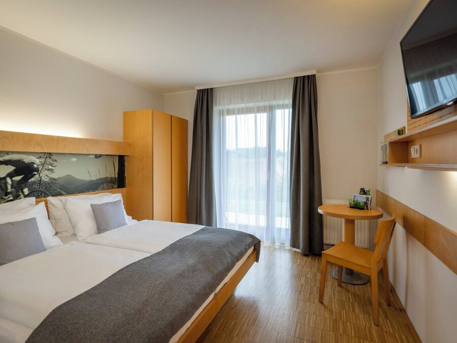 Ein Hotelzimmer mit Bett, kleinem Tisch, Stuhl, Fernseher und einem Fenster mit Vorhängen.