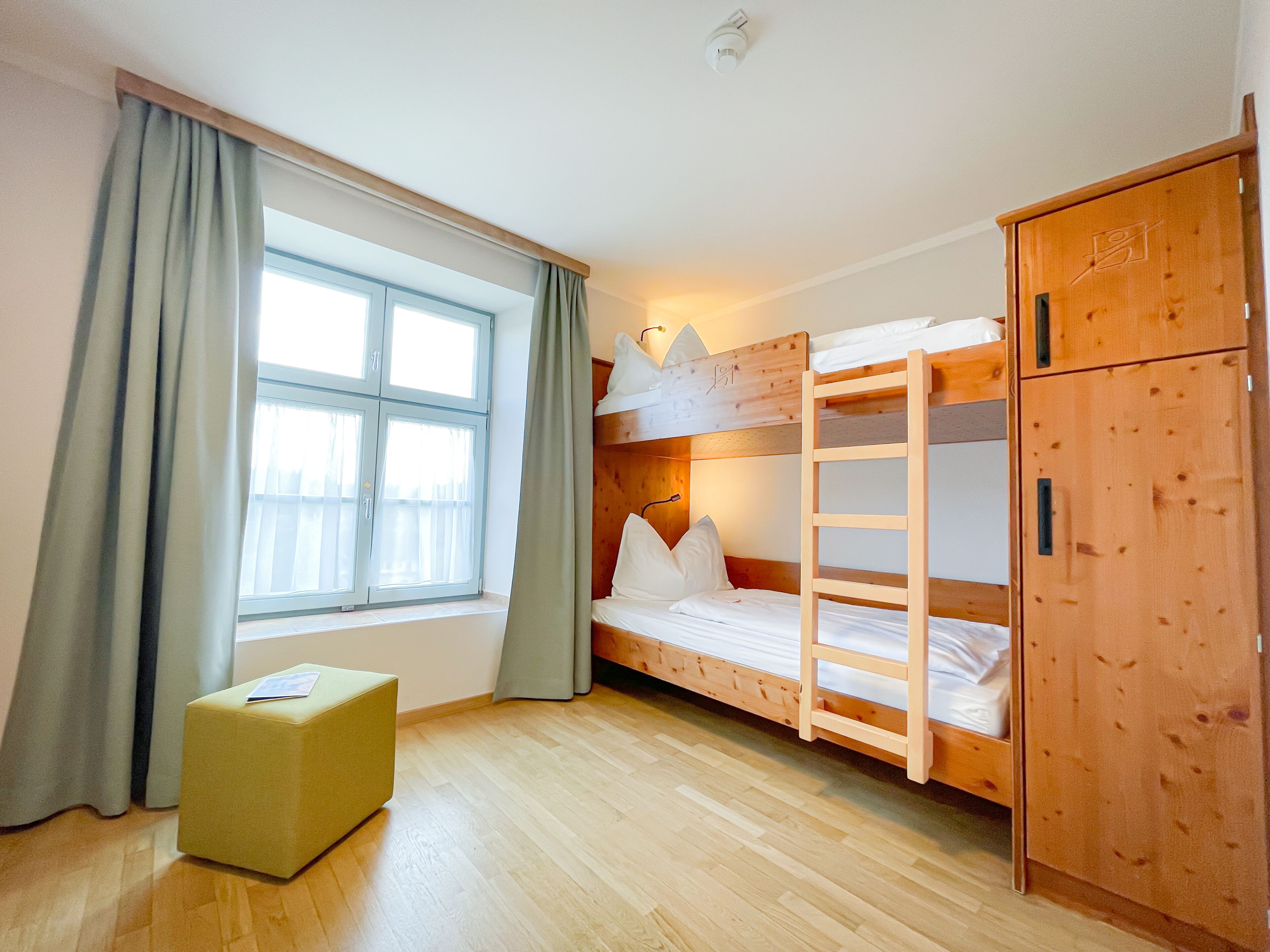 A bunk bed room with a window, curtains, a wooden wardrobe, and a small stool.