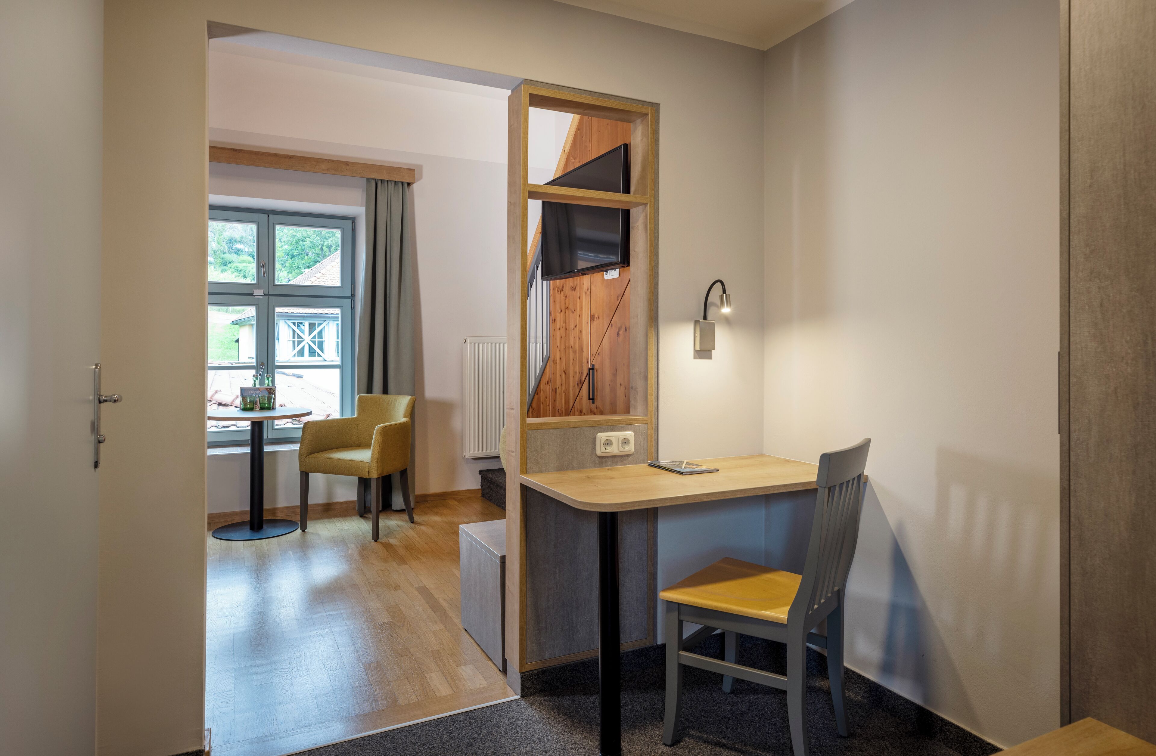 Una habitación pequeña con un escritorio de madera, una silla, una mesita con otra silla, y una ventana con cortinas.