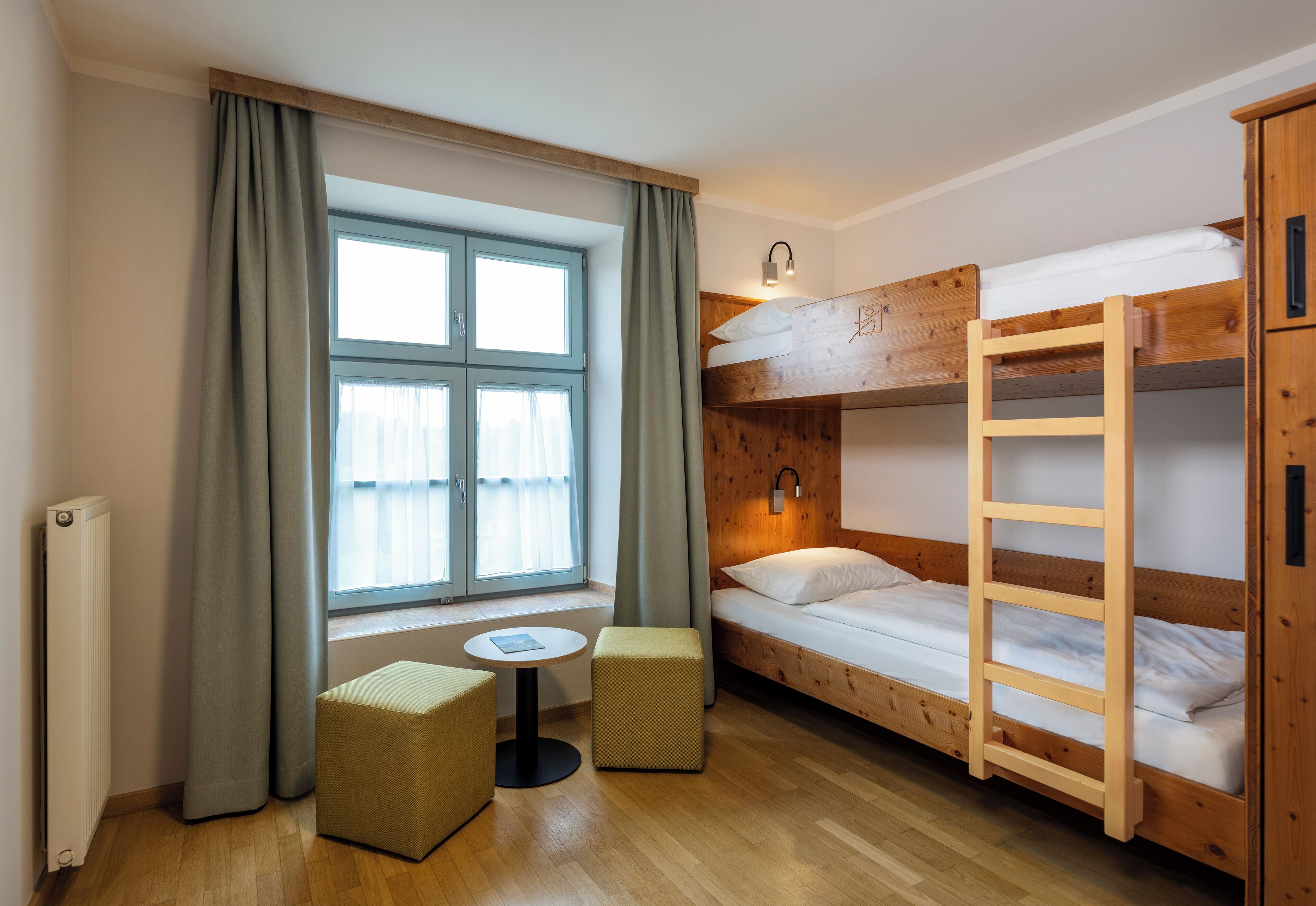 Een kleine kamer met een stapelbed, een tafeltje, twee voetenbankjes en een raam met gordijnen.