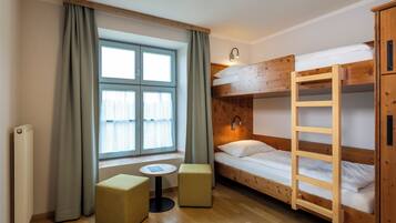Een kleine kamer met een stapelbed, een tafeltje, twee voetenbankjes en een raam met gordijnen.