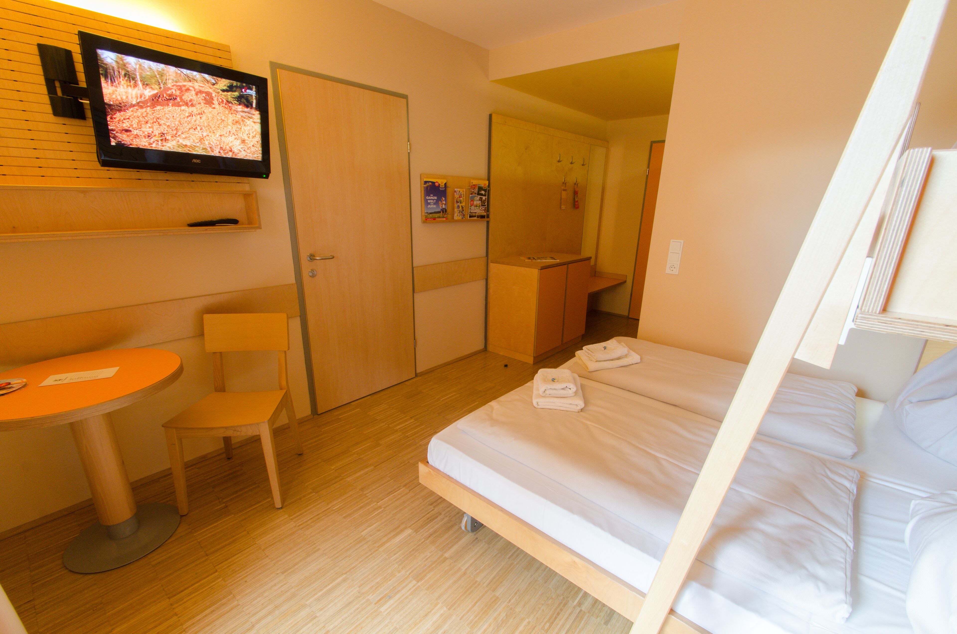 A hotel room with a single bed, a small table, a chair, a TV mounted on the wall, and a wooden wardrobe.