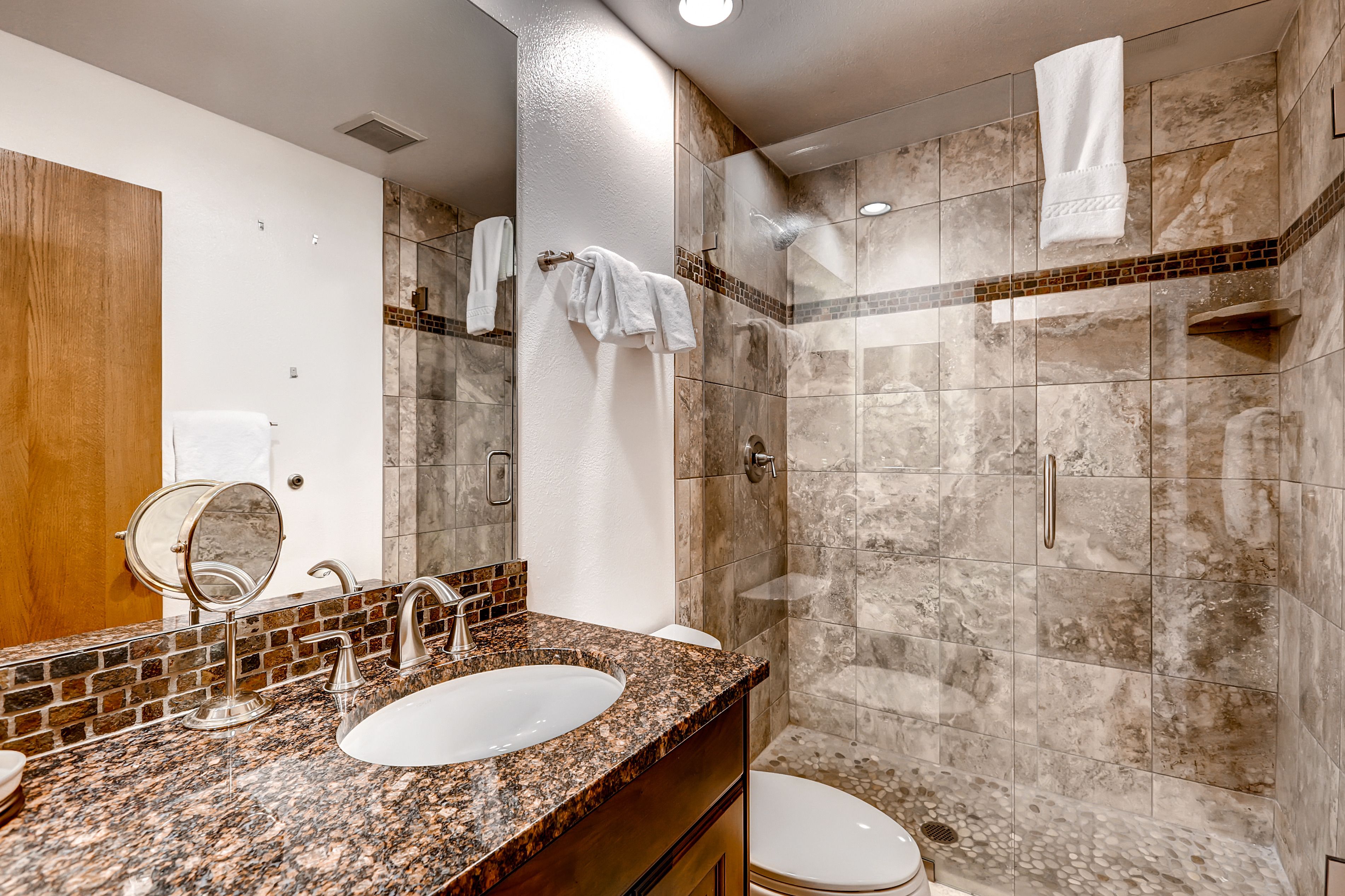 Un baño con encimera de granito, dos lavamanos, un espejo y zona de ducha con paredes de azulejos.