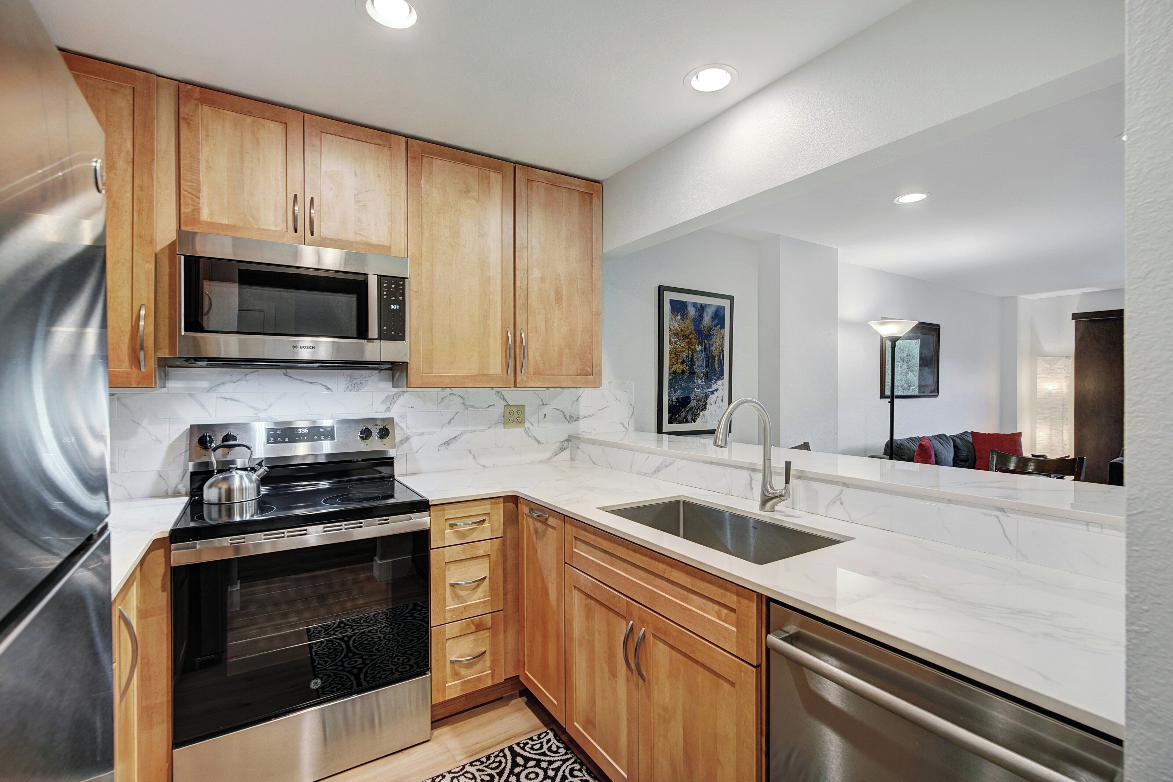 A modern kitchen with wooden cabinets, stainless steel appliances, a marble countertop, and a built-in oven.
