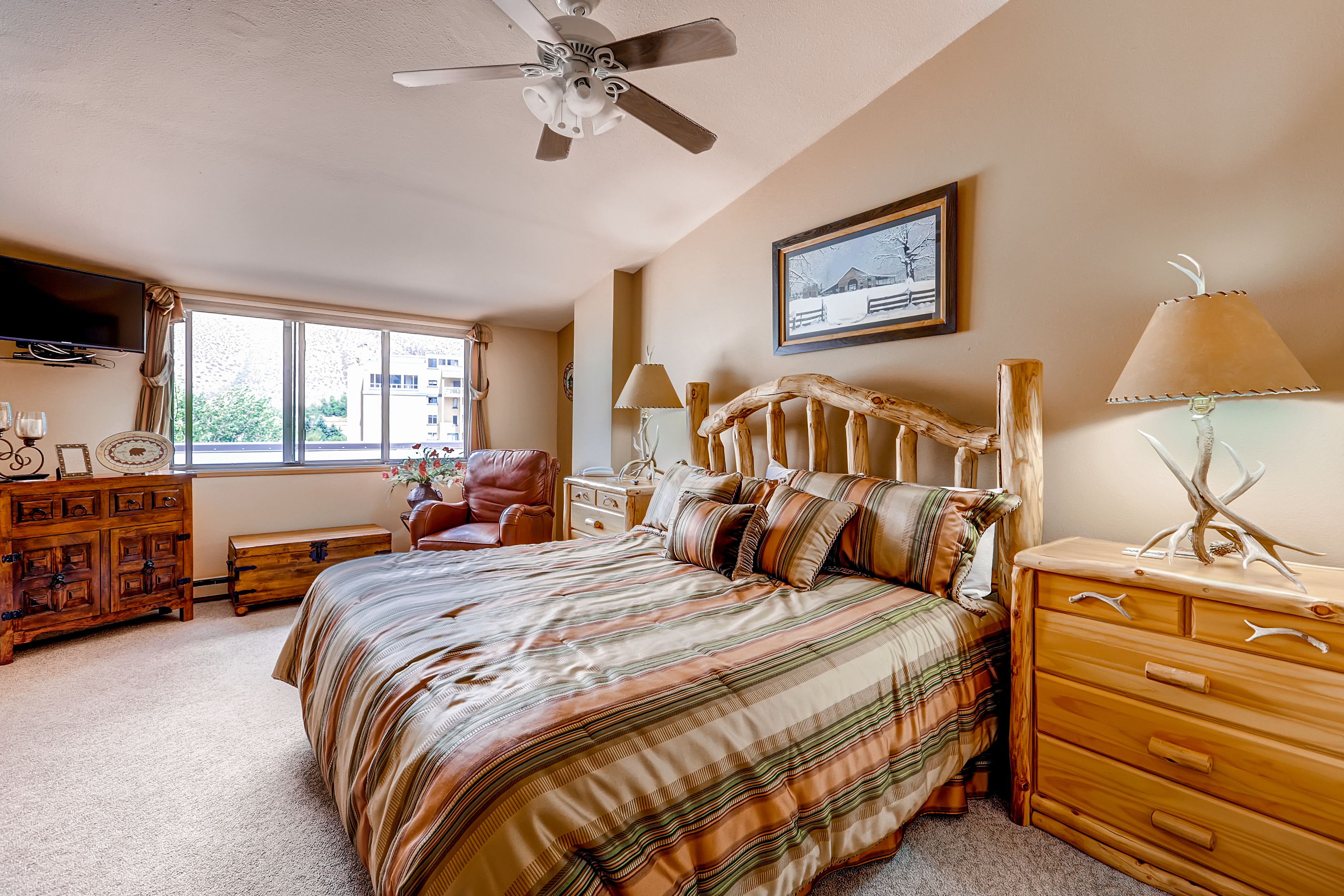 A bedroom with a bed, wooden dresser, nightstands, a ceiling fan, a window with a view, and a framed picture on the wall.