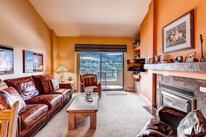 A living room with a leather sofa, a coffee table, and a fireplace. There is a balcony with a view of the countryside.