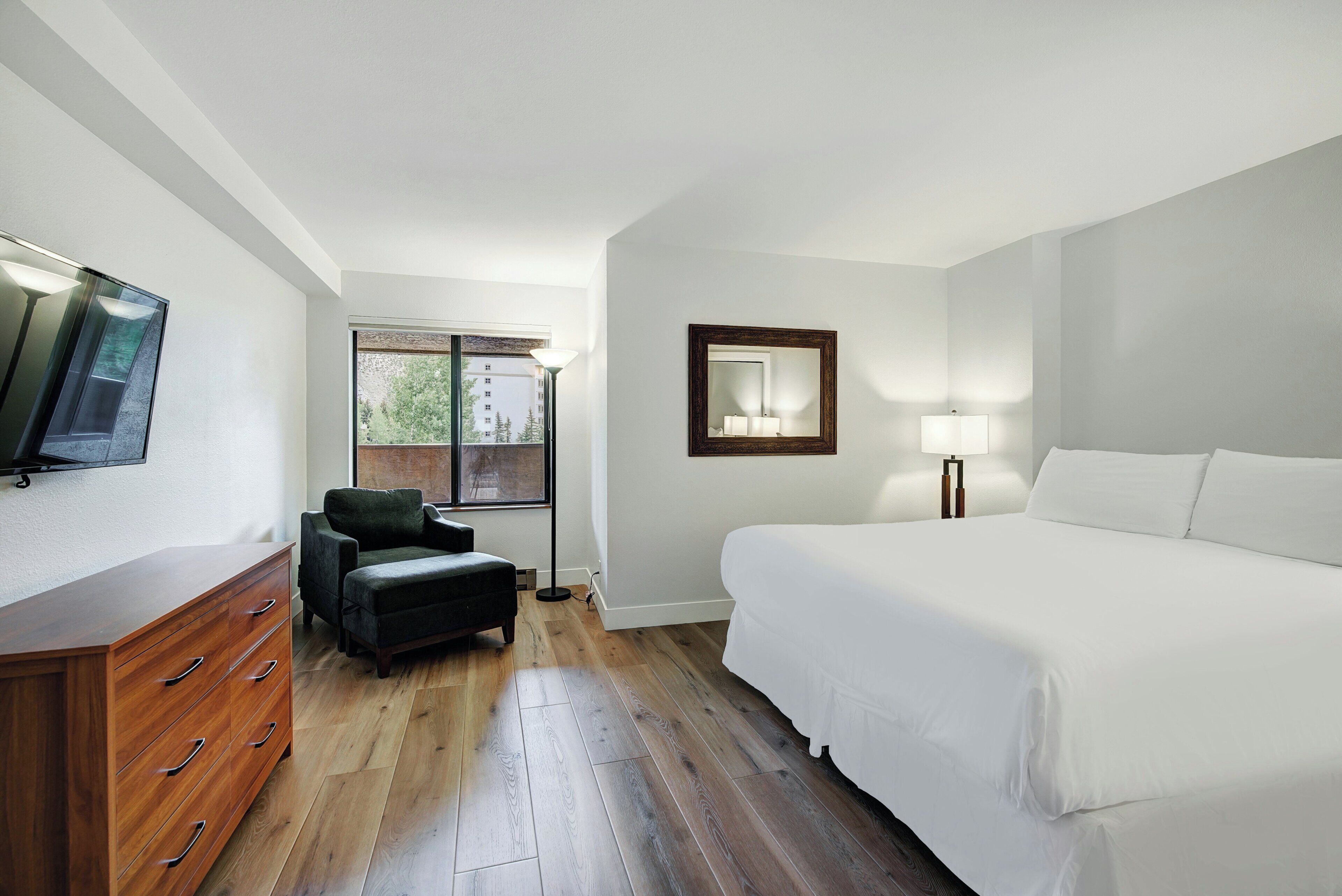 A hotel room with a bed, a dresser, a chair, a TV, a window, and a lamp.