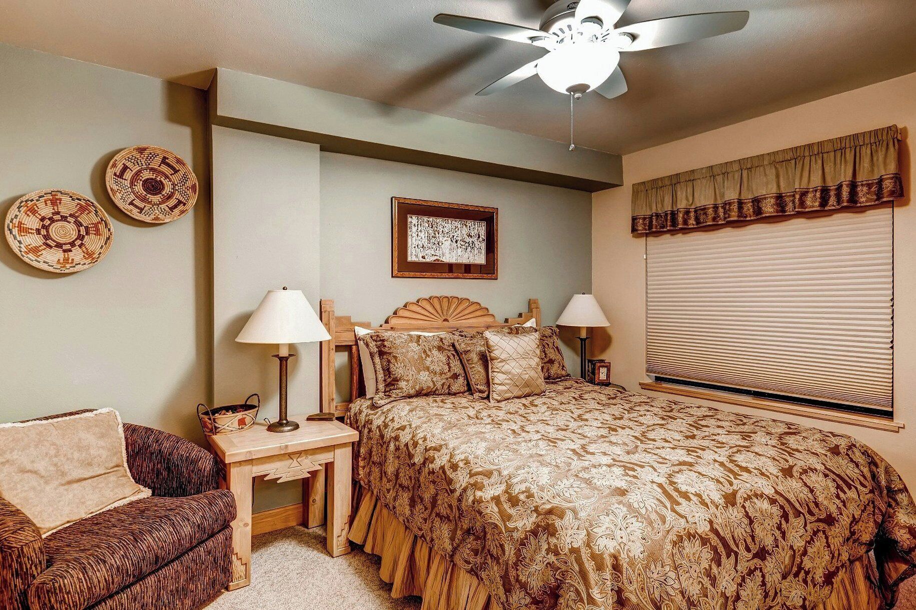 A bedroom with a bed, two bedside tables, a chair, a ceiling fan, a window with blinds, and wall decor.