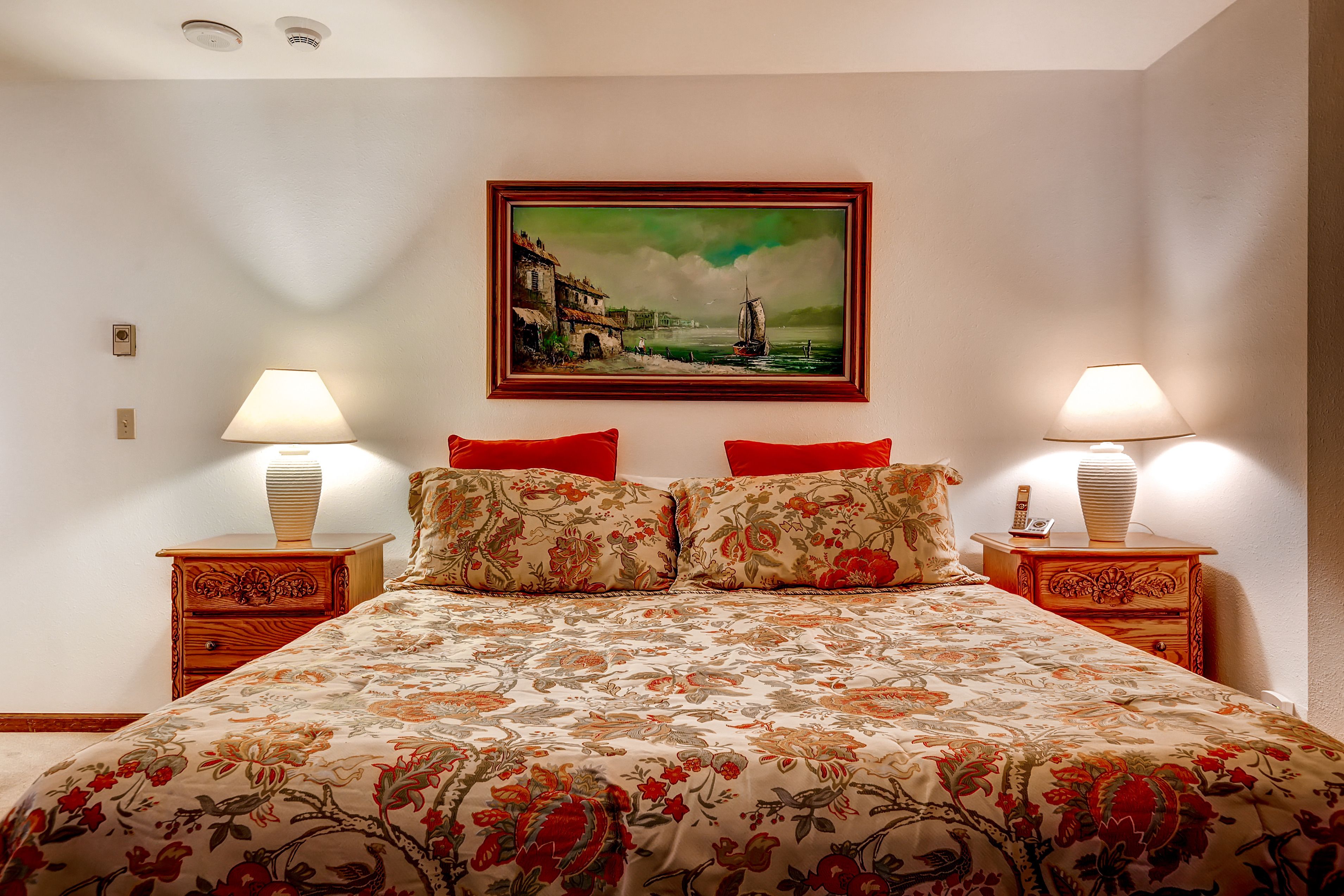 Hotelkamer met een bed, bloemenprint sprei, twee nachtkastjes met lampen en een lijst met schilderij aan de muur.