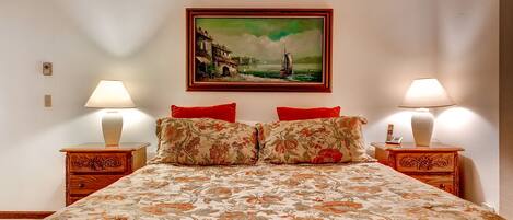 Hotelkamer met een bed, bloemenprint sprei, twee nachtkastjes met lampen en een lijst met schilderij aan de muur.