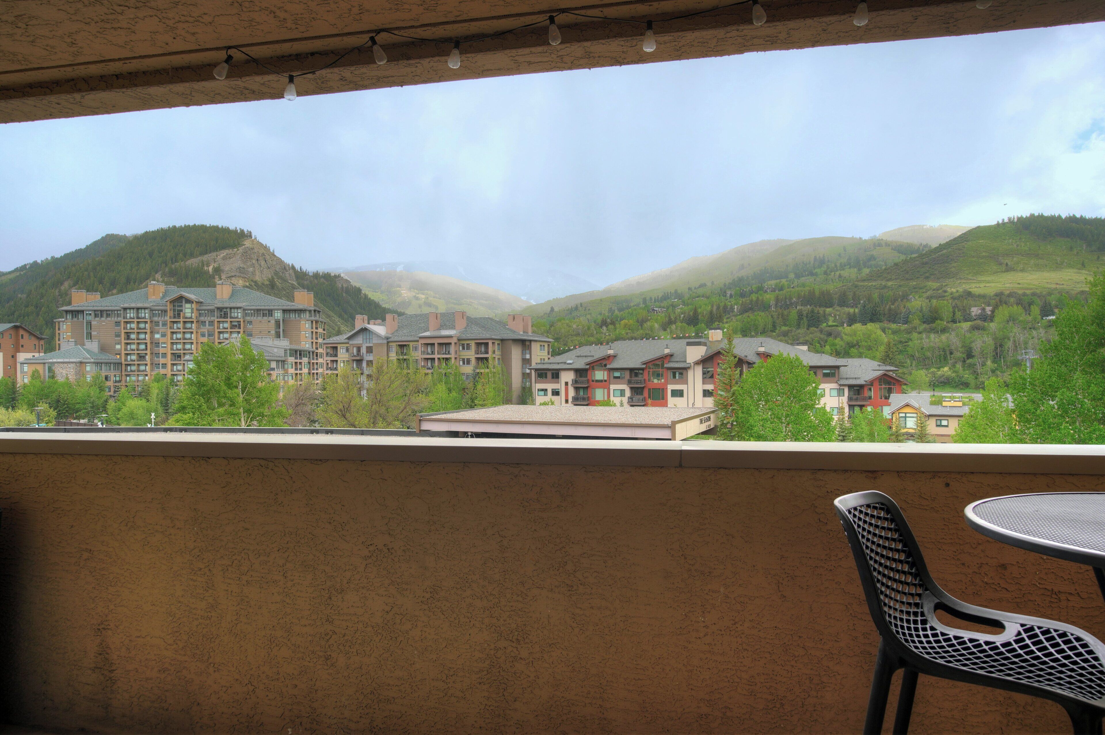 A balcony with a view of a mountain, a building with multiple floors, and a table with a chair.